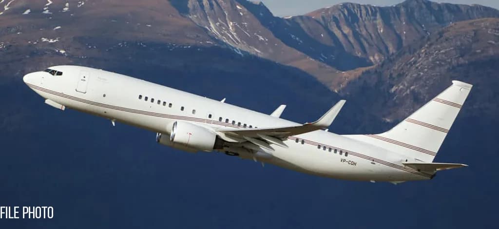 A white Boeing 737-style business jet with red pinstripes climbs against a backdrop of mountains.