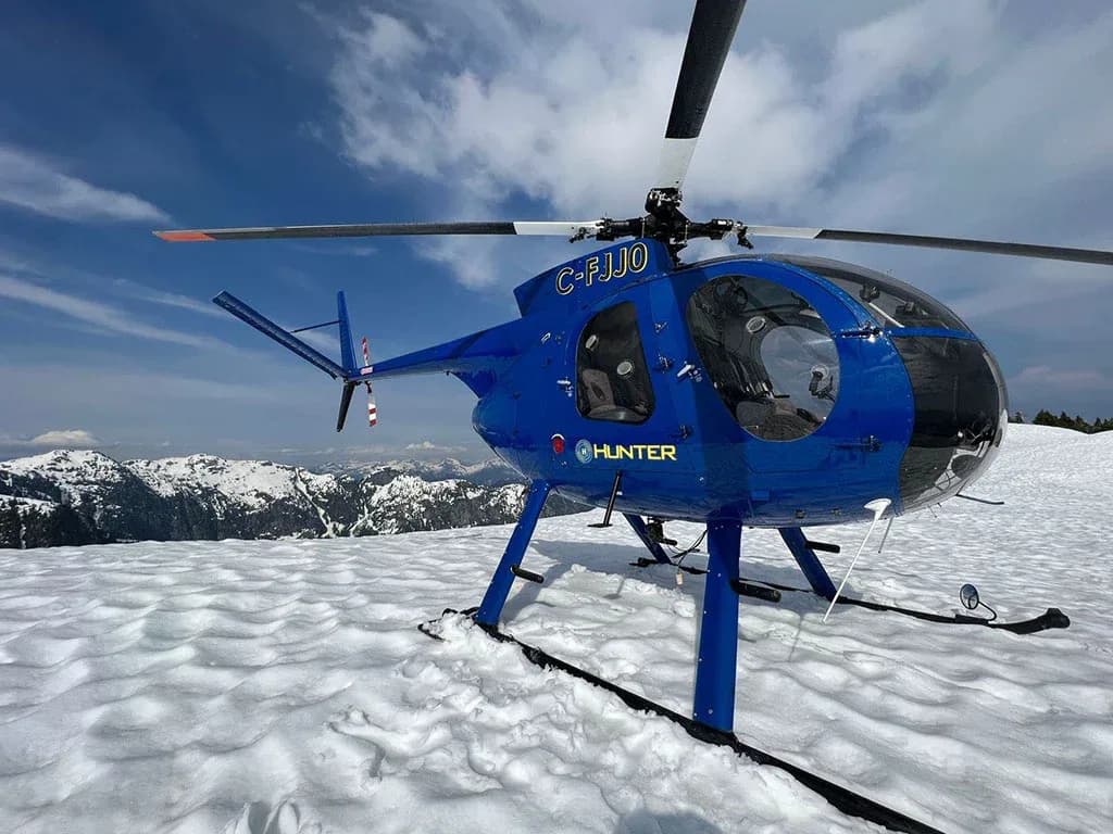 A blue helicopter with registration C-FJJ0 sits on a snowy mountain peak under a blue sky.
