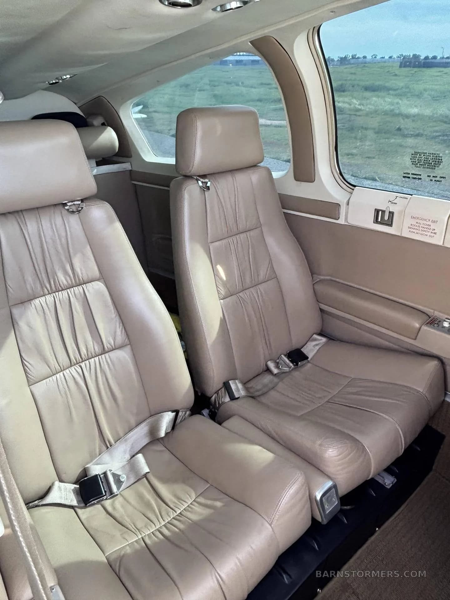 The interior cabin of a small aircraft, showing two tan leather passenger seats with seatbelts and a view of green fields outside the window.
