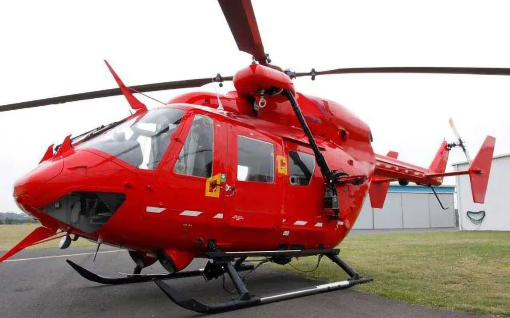 A red helicopter with skids parked on an asphalt surface in front of a building.