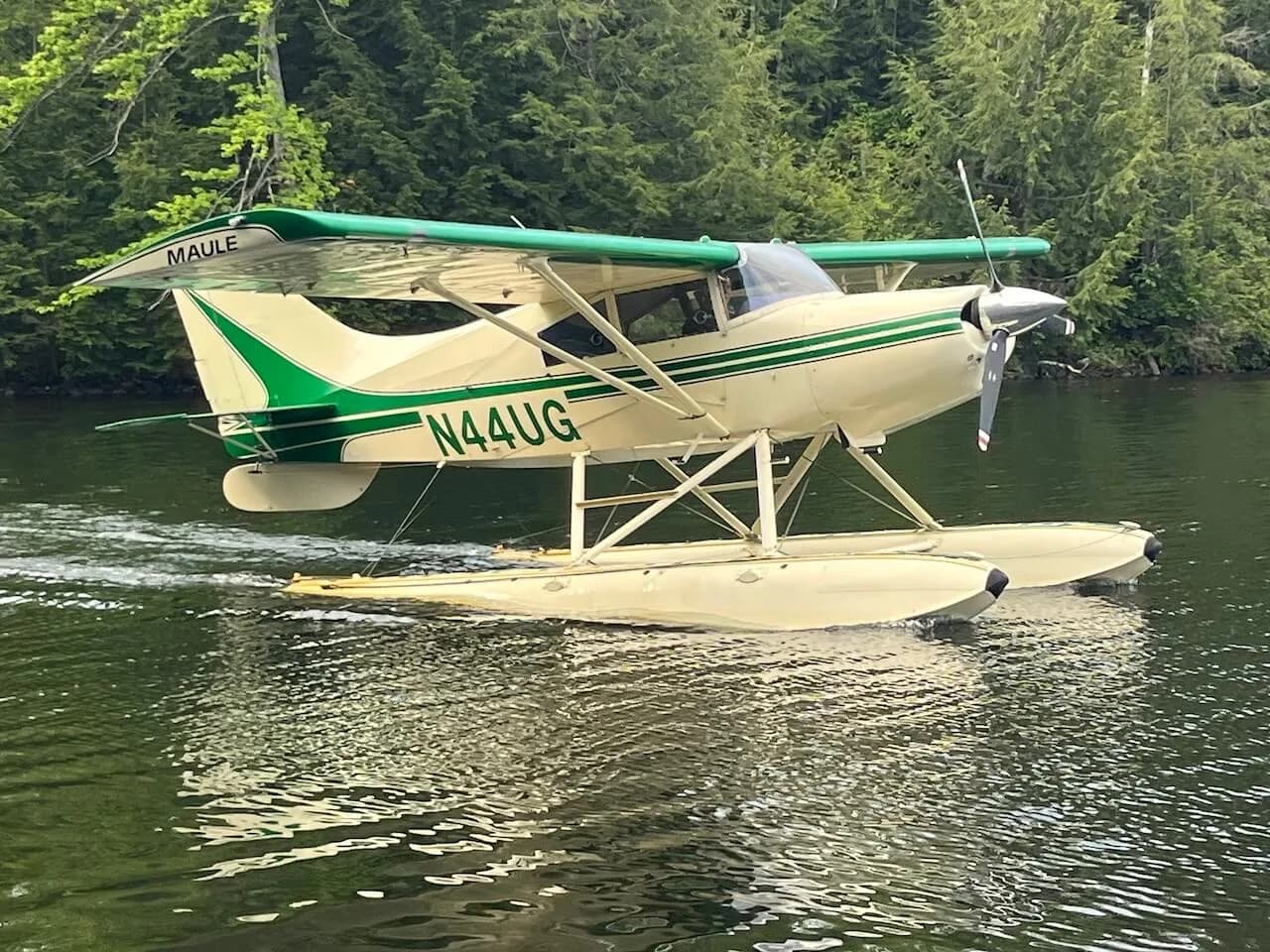 A Maule seaplane with a green and white paint scheme on floats is moving across a lake.
