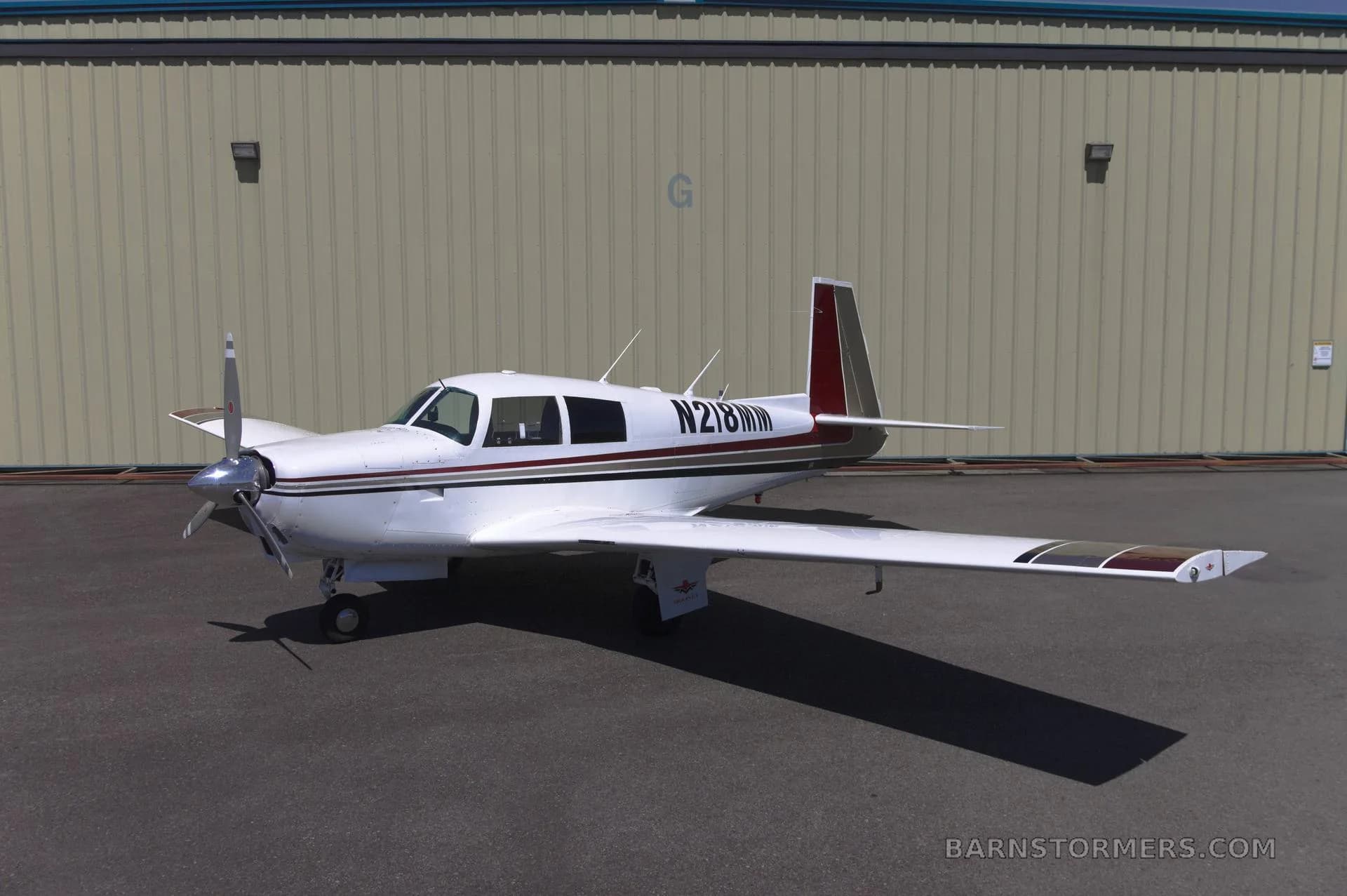 A white single-engine propeller aircraft with red and brown stripes, viewed from the front-left, parked on an asphalt surface in front of a light-colored hangar.