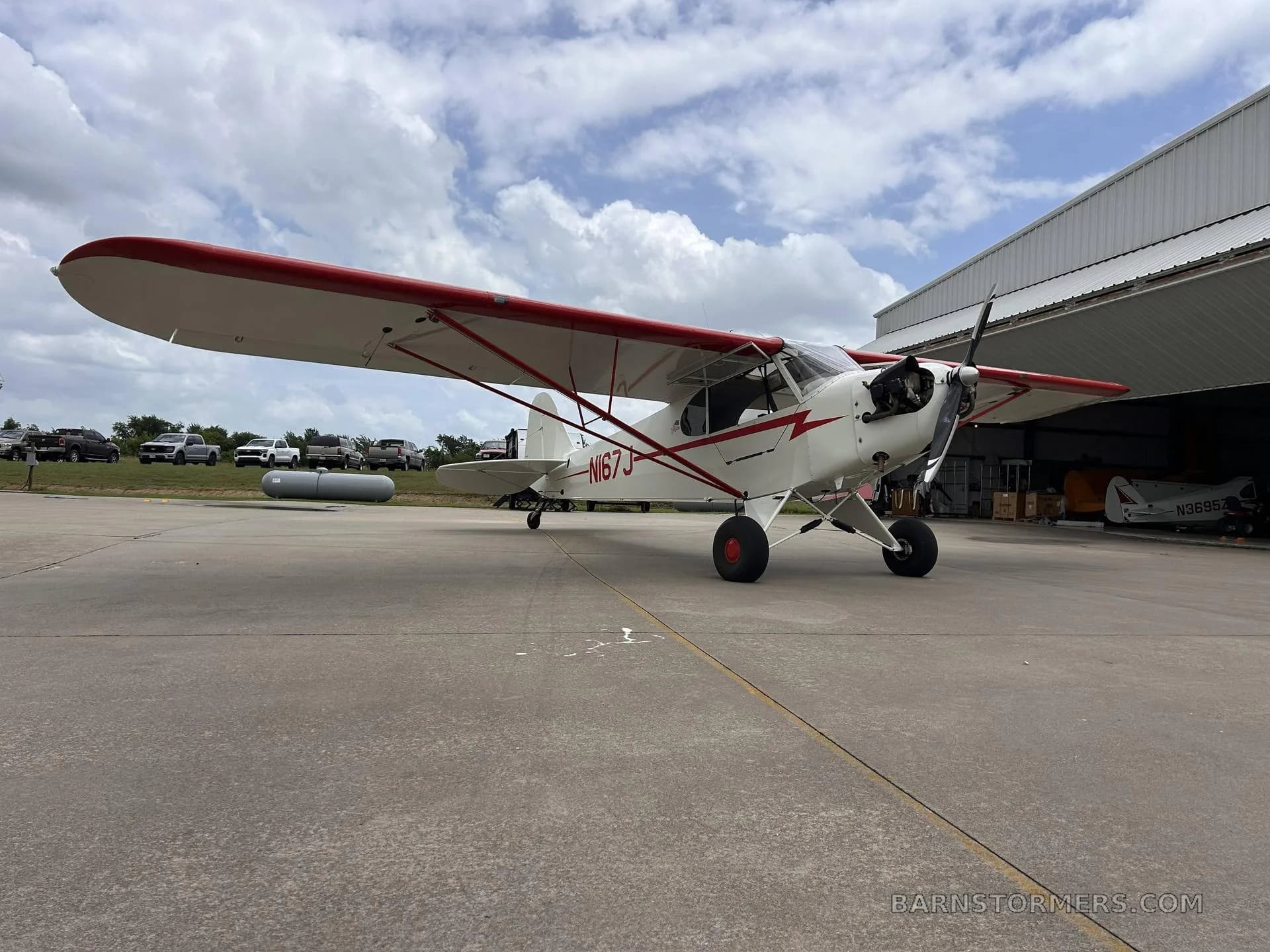 A low-angle view of a white and red single-engine aircraft (N167J) parked on an airfield with hangars and vehicles in the background.