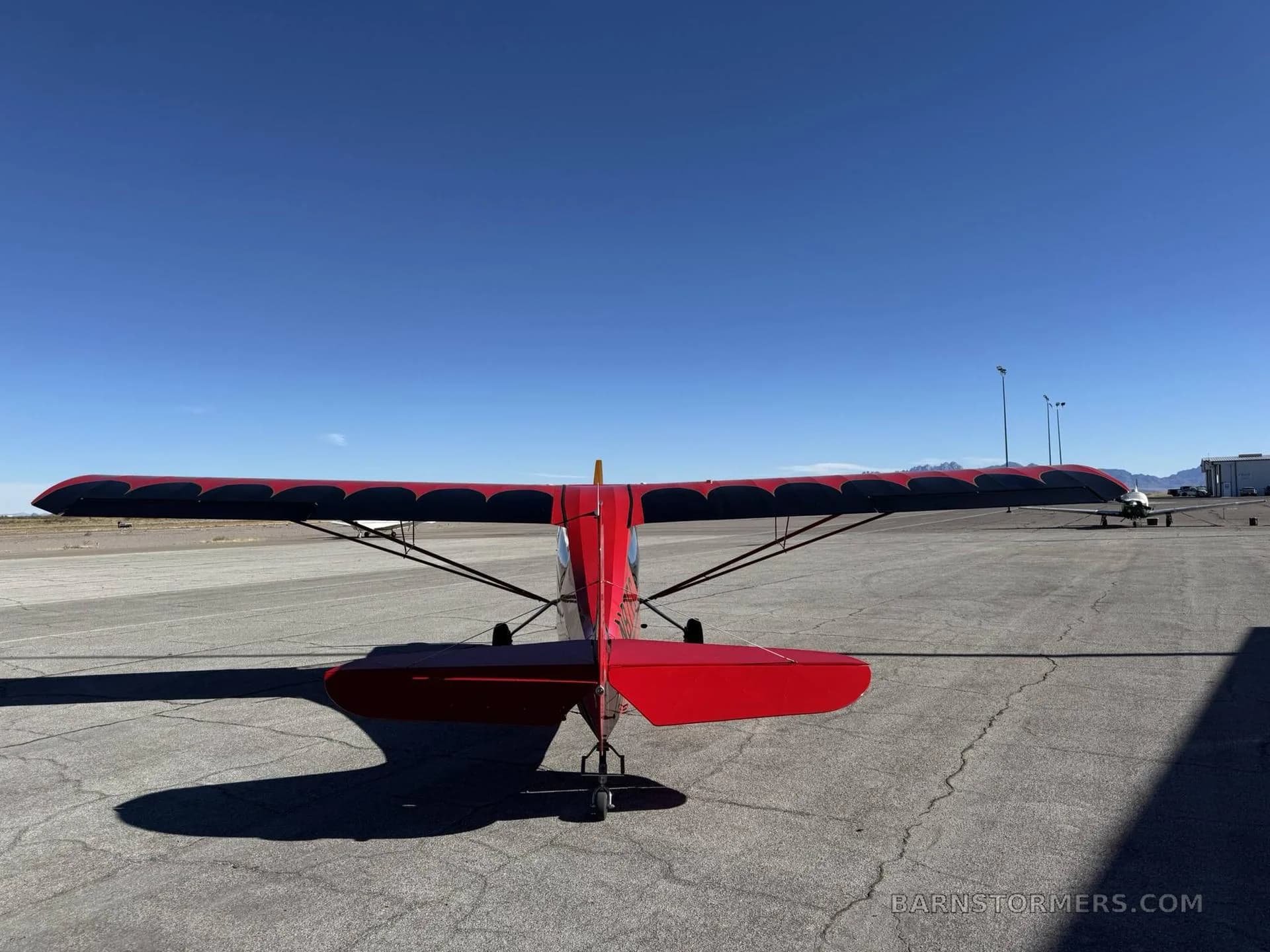A red high-wing monoplane with black wing accents is viewed from the rear, parked on an airfield under a clear blue sky.