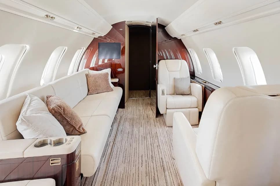 A view inside the aircraft cabin shows a long cream leather sofa with throw pillows opposite a single armchair, with dark wood paneling at the rear.