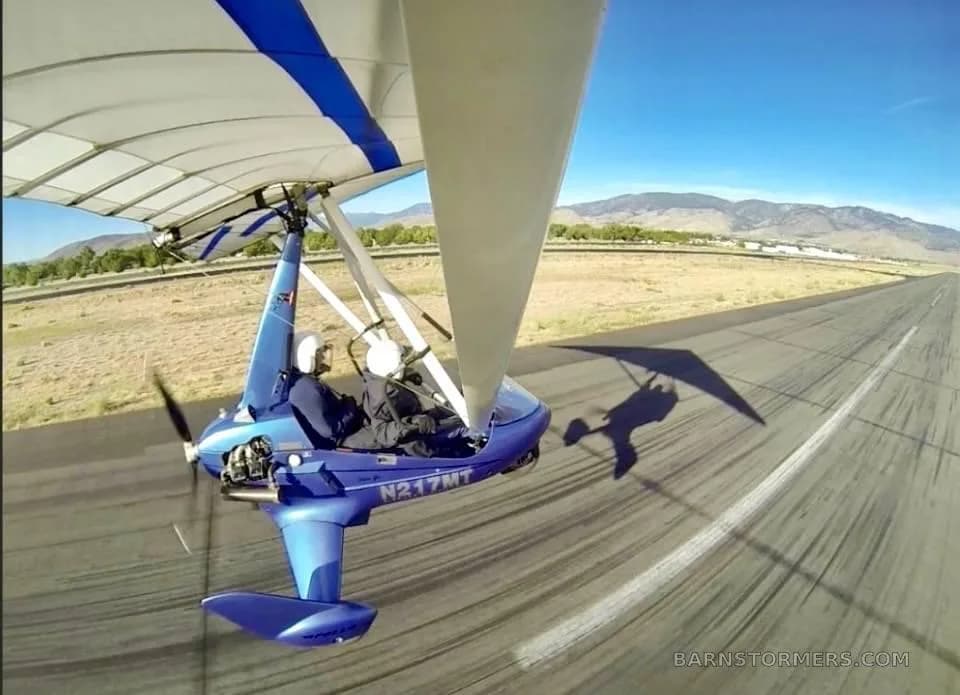 A blue ultralight aircraft with registration N217MT is seen from above, flying low over a runway with a pilot and passenger.