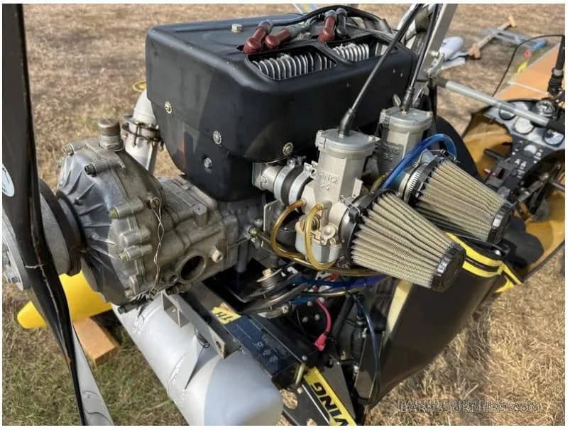 A close-up view of the engine and propeller of a yellow ultralight aircraft, showing the air filters and mechanical components.