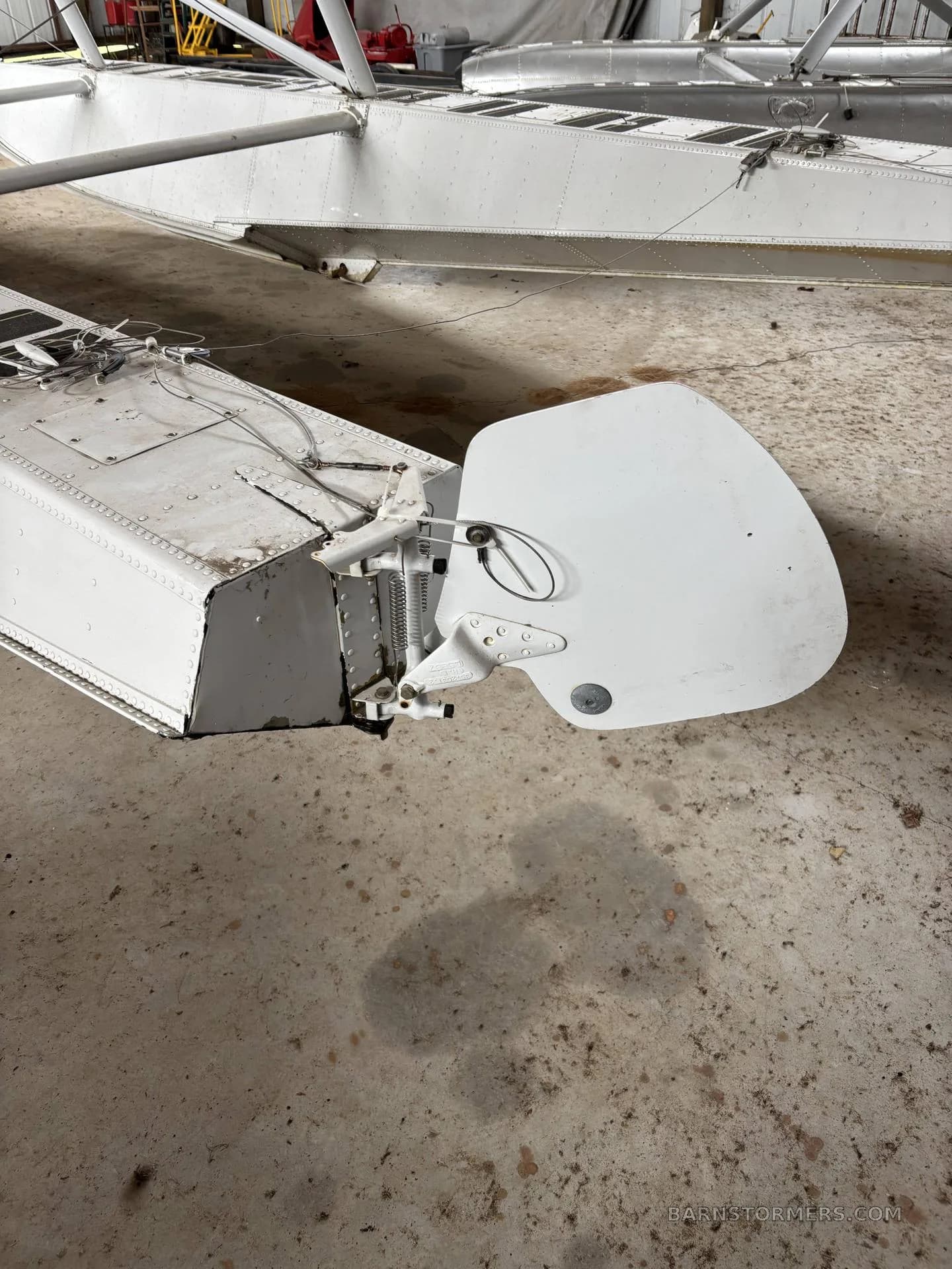A close-up of the rear section of a white aircraft float with its rudder assembly, showing control cables and rivets, on a concrete floor.