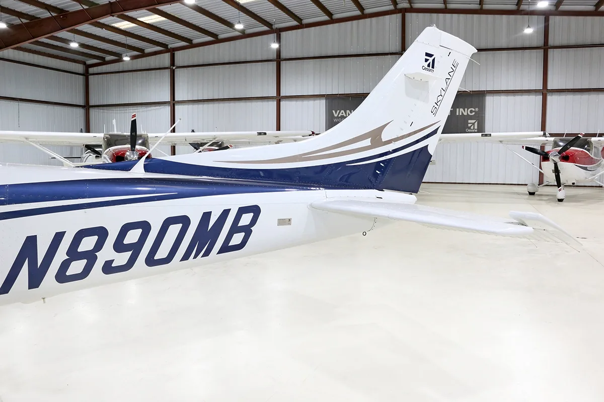 The tail section of a white and blue Cessna Skylane N890MB is shown in a hangar.