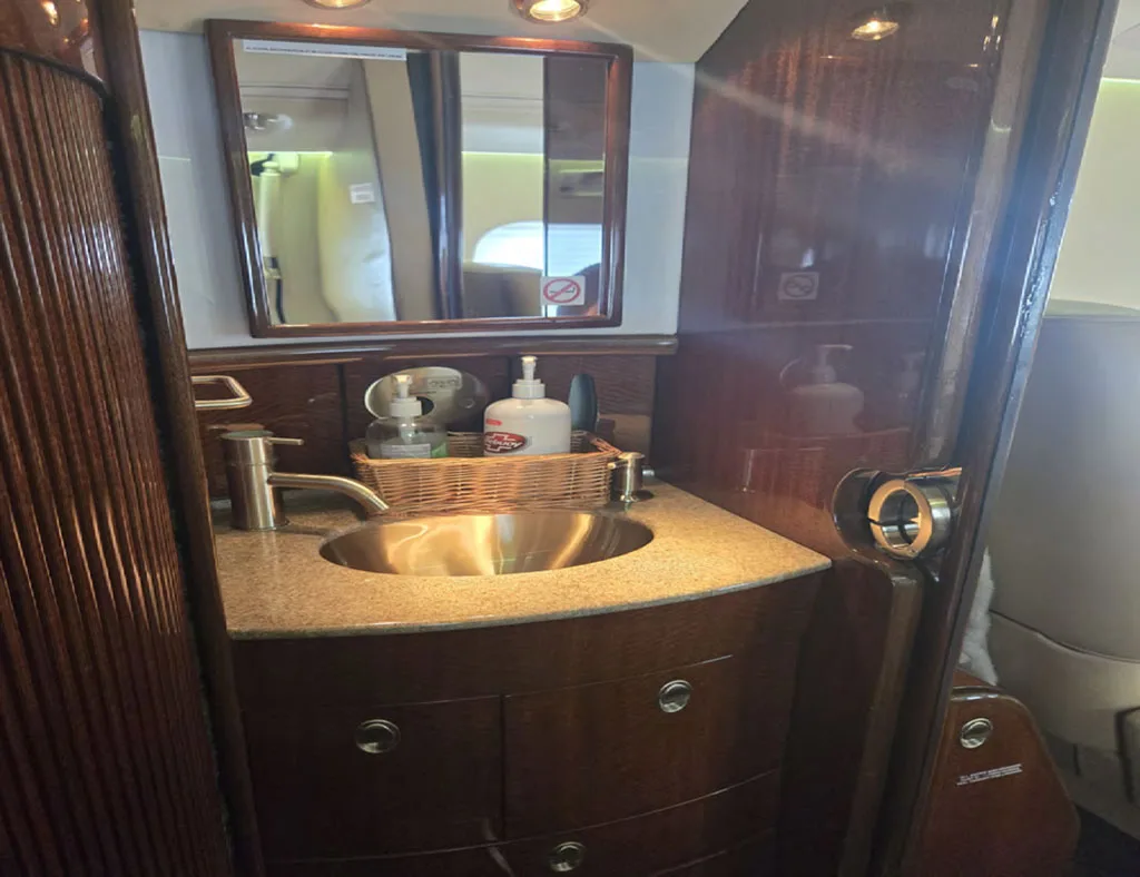 A small lavatory or galley area inside a business jet, featuring a sink, mirror, and wood cabinetry.