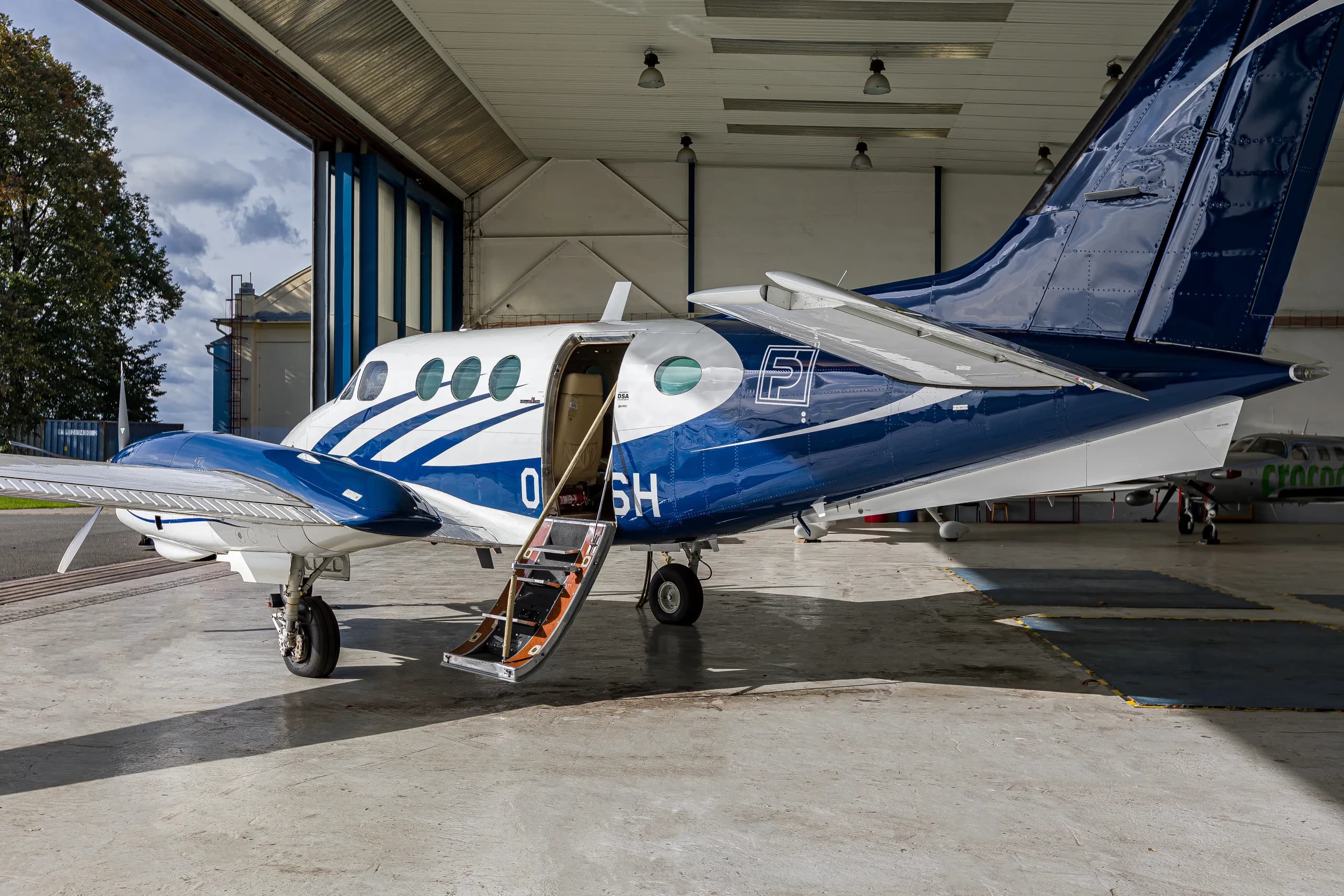 A white and blue twin-engine turboprop aircraft, registration OK-DSH, with its passenger door open and stairs extended, parked in a hangar.