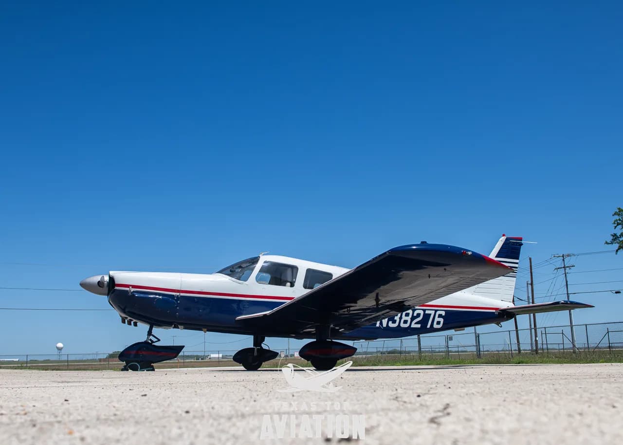A side view of the Piper aircraft N38276, painted white with blue and red stripes, parked on the tarmac.