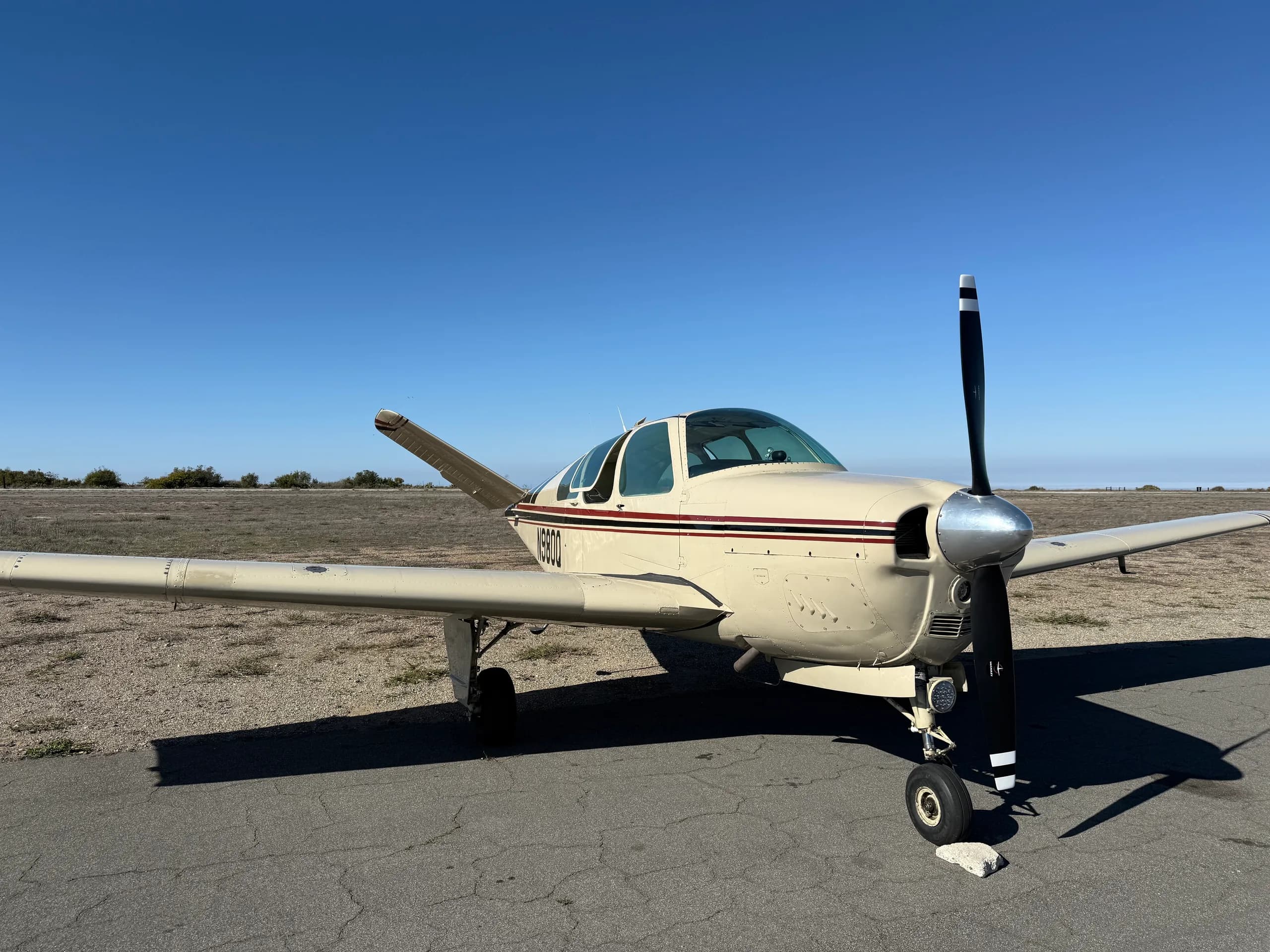 A cream-colored single-engine propeller aircraft with red and black stripes is parked on an asphalt ramp under a clear blue sky.