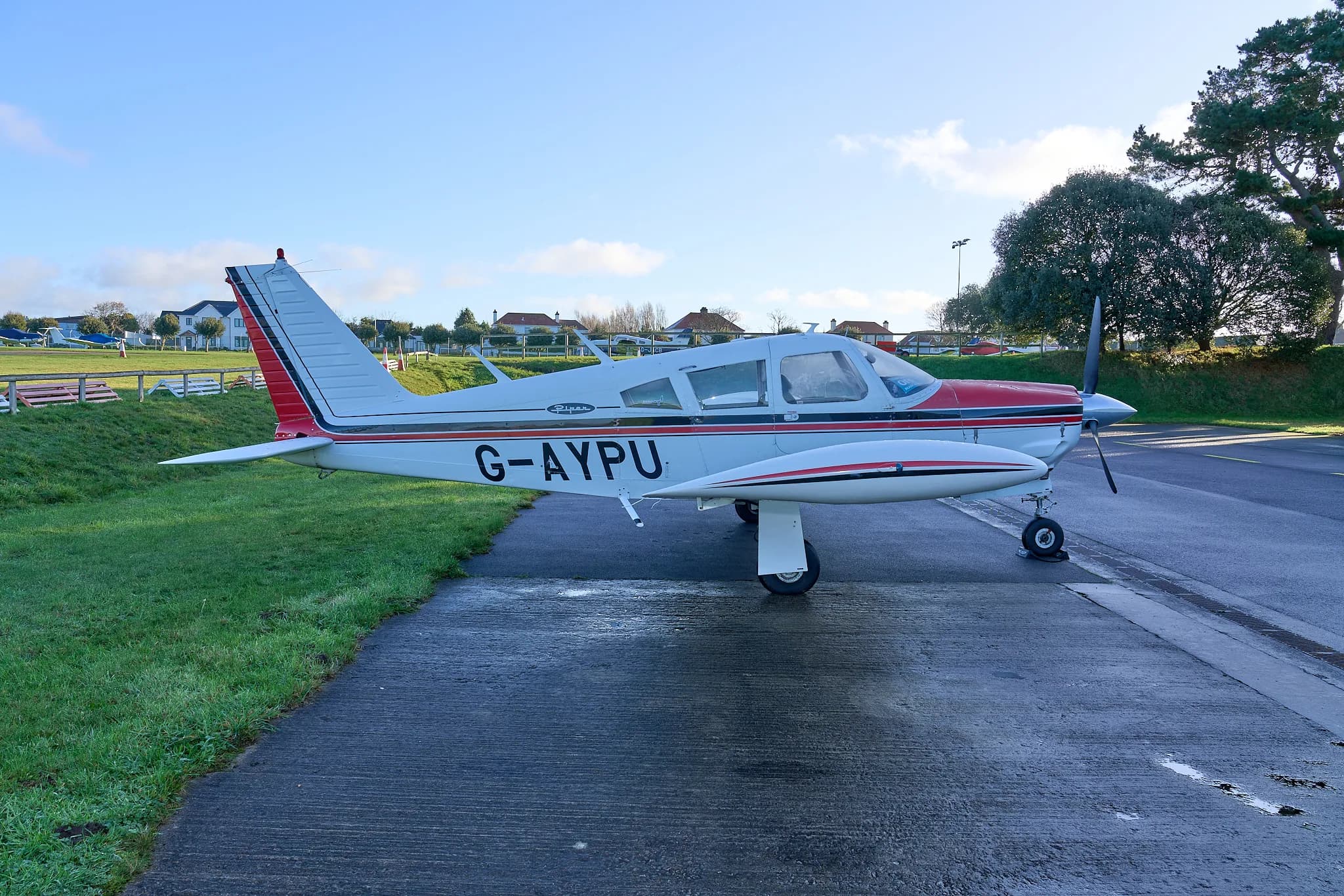 A side view of the white and red Piper Cherokee G-AYPU on the tarmac with houses in the distance.