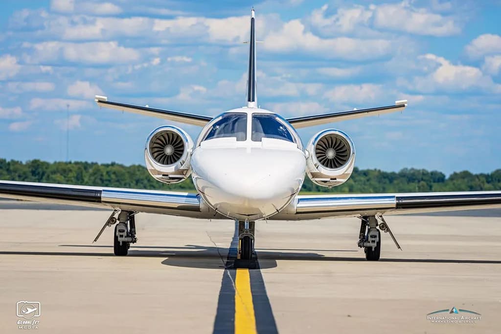 A head-on view of a white Cessna Citation Bravo business jet with red and black stripes on an airport tarmac.