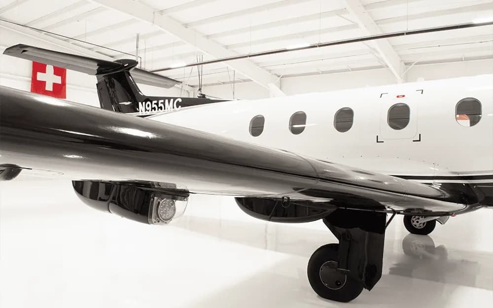Close-up of the black wing, white fuselage, and landing gear of a Pilatus PC-12 aircraft in a hangar.