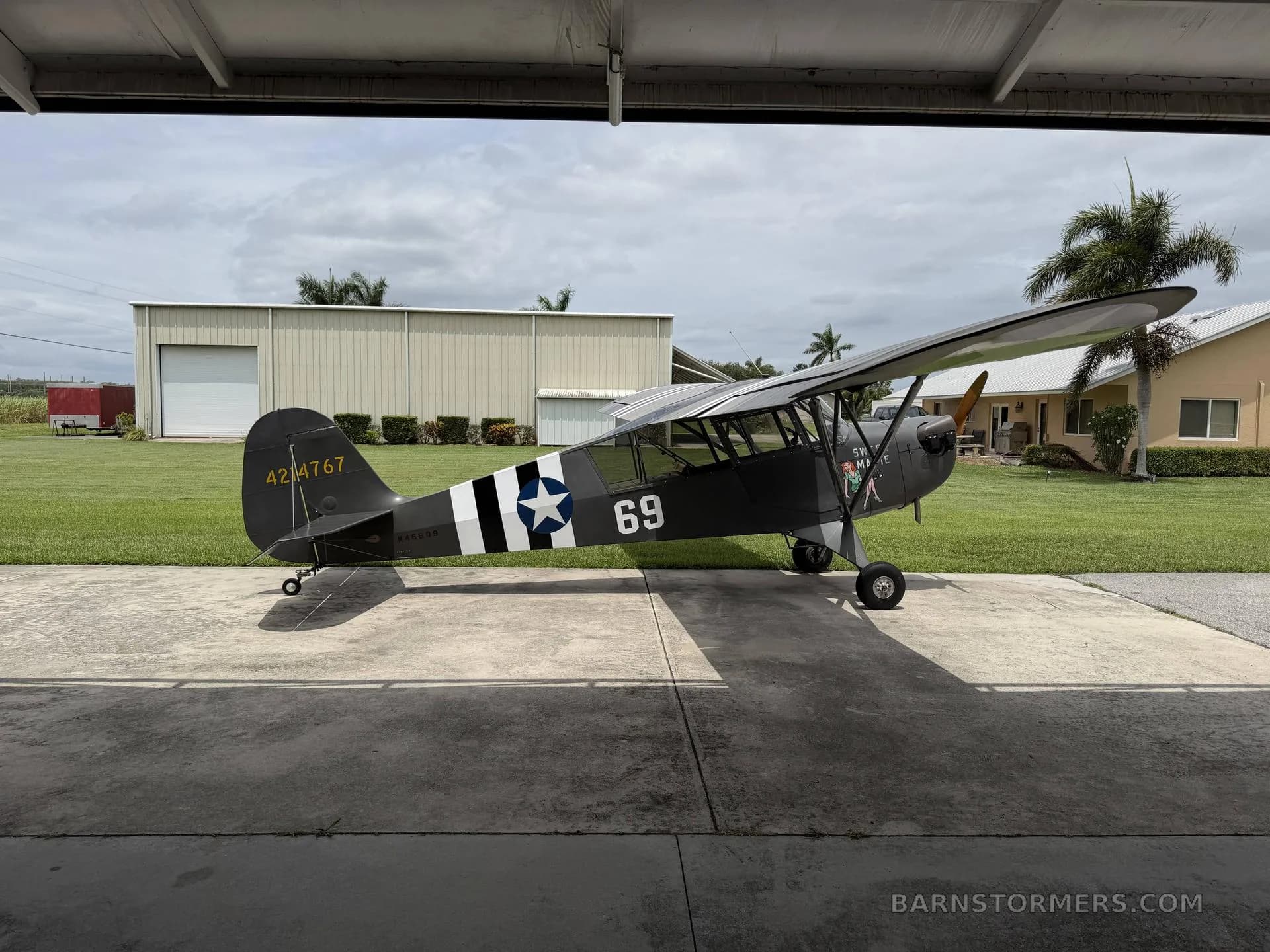 A side view of a dark green Piper Cub-style aircraft with military markings, parked on concrete under a cloudy sky.