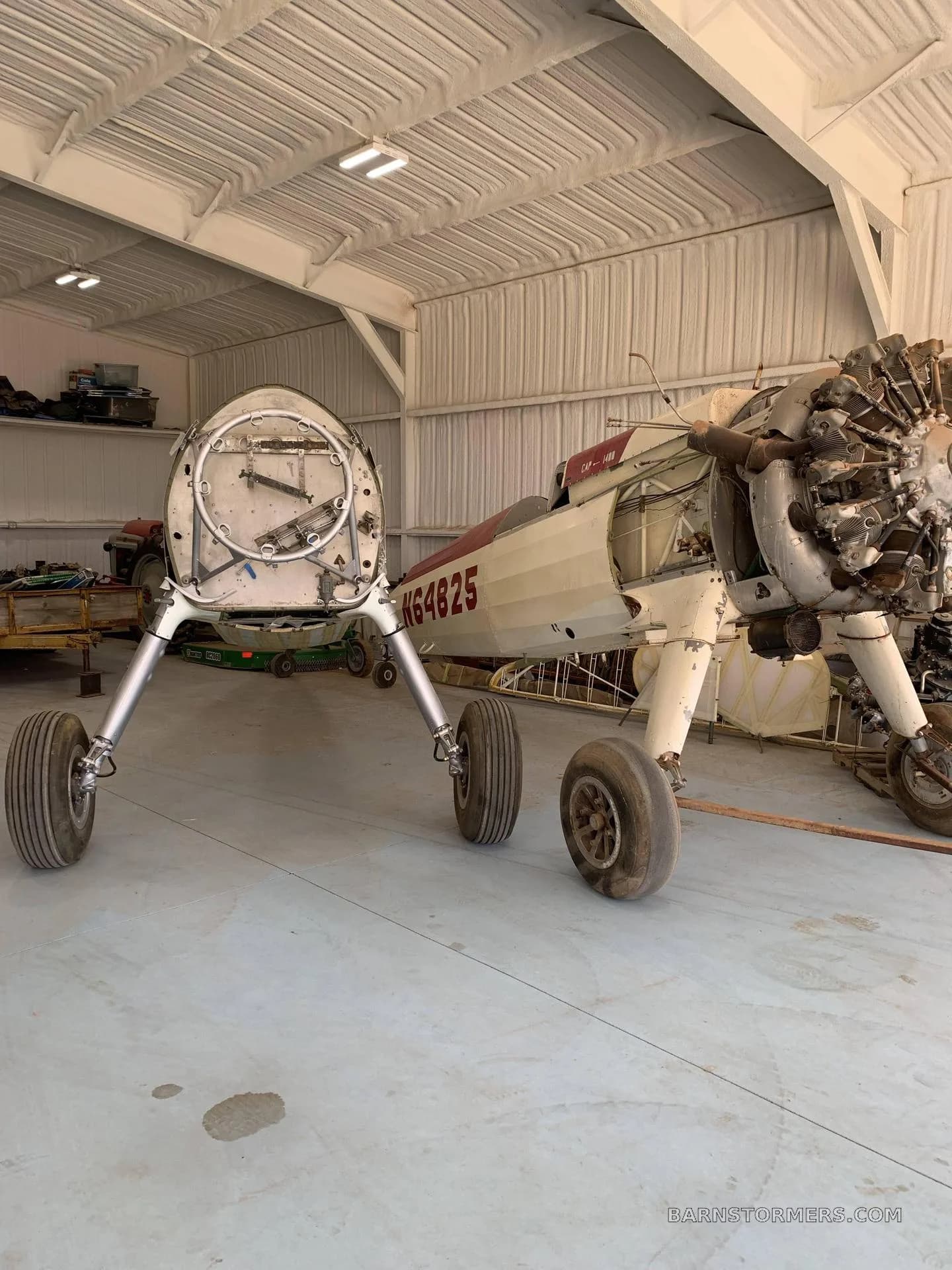 A partially disassembled white and red biplane fuselage with a radial engine and landing gear, inside a hangar.