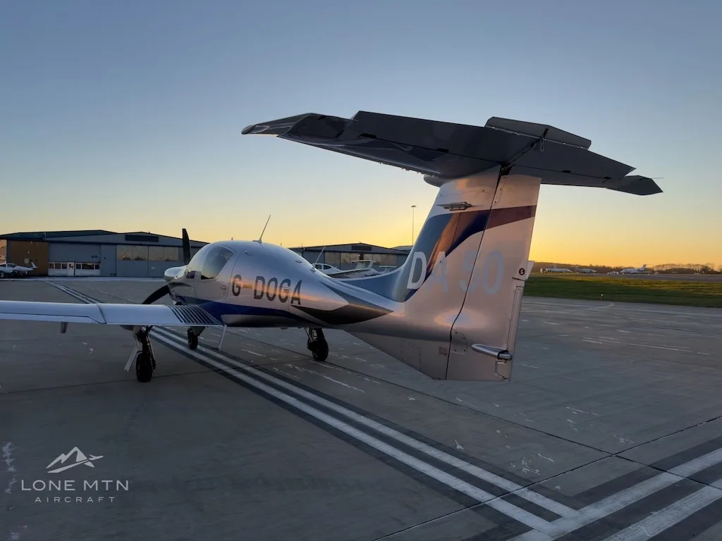 A rear quarter view of a silver and blue Diamond DA50 aircraft with registration G-DOGA parked on an airfield at sunset.