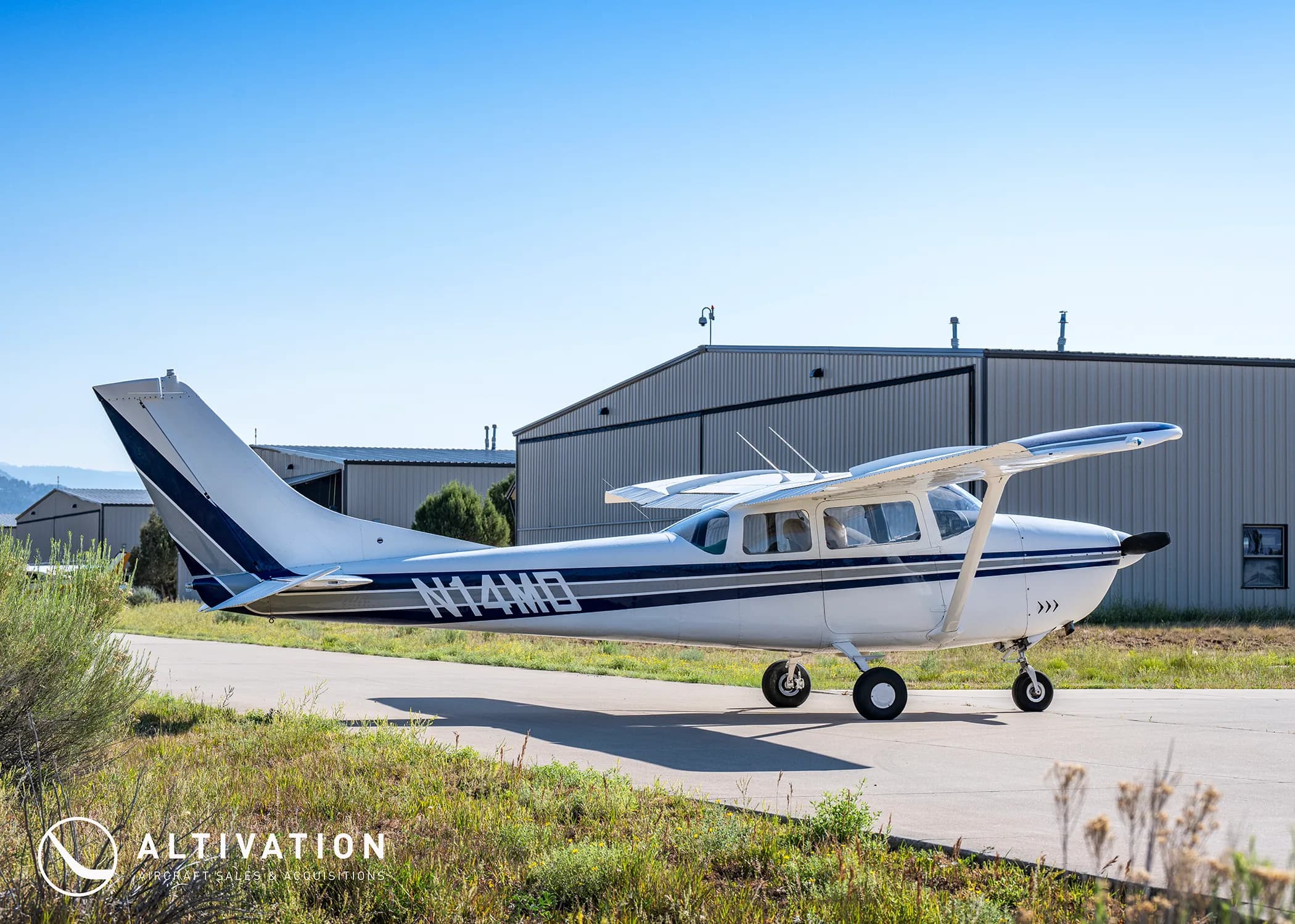 A white Cessna 182 with blue and grey stripes, registration N74MD, parked on a tarmac in front of hangars.