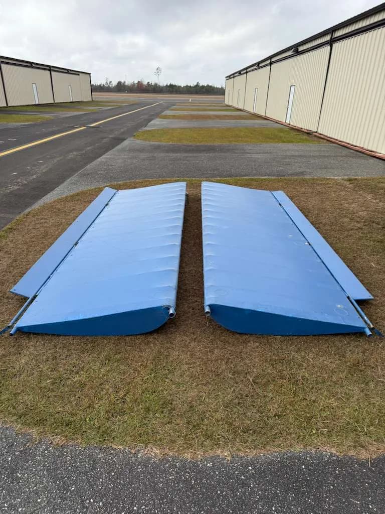 Two detached blue aircraft wings lying on the grass next to an airfield runway.