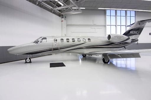 A silver Cessna Citation CJ3+ aircraft with black and grey stripes on the fuselage and tail, parked inside a brightly lit hangar.