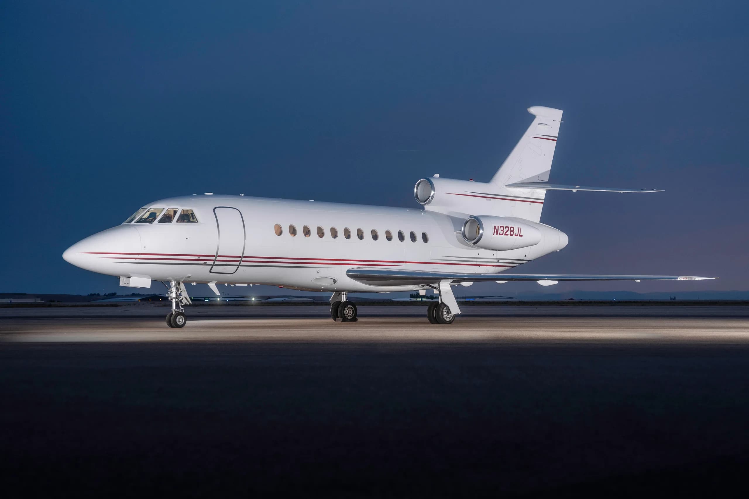 A white Dassault Falcon business jet with red pinstripes is parked on an airfield at dusk.