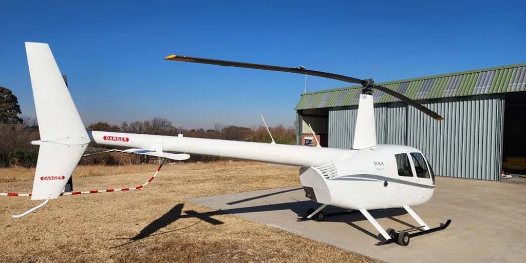 A white Robinson R44 helicopter is parked on a concrete pad with a green hangar and trees in the background.
