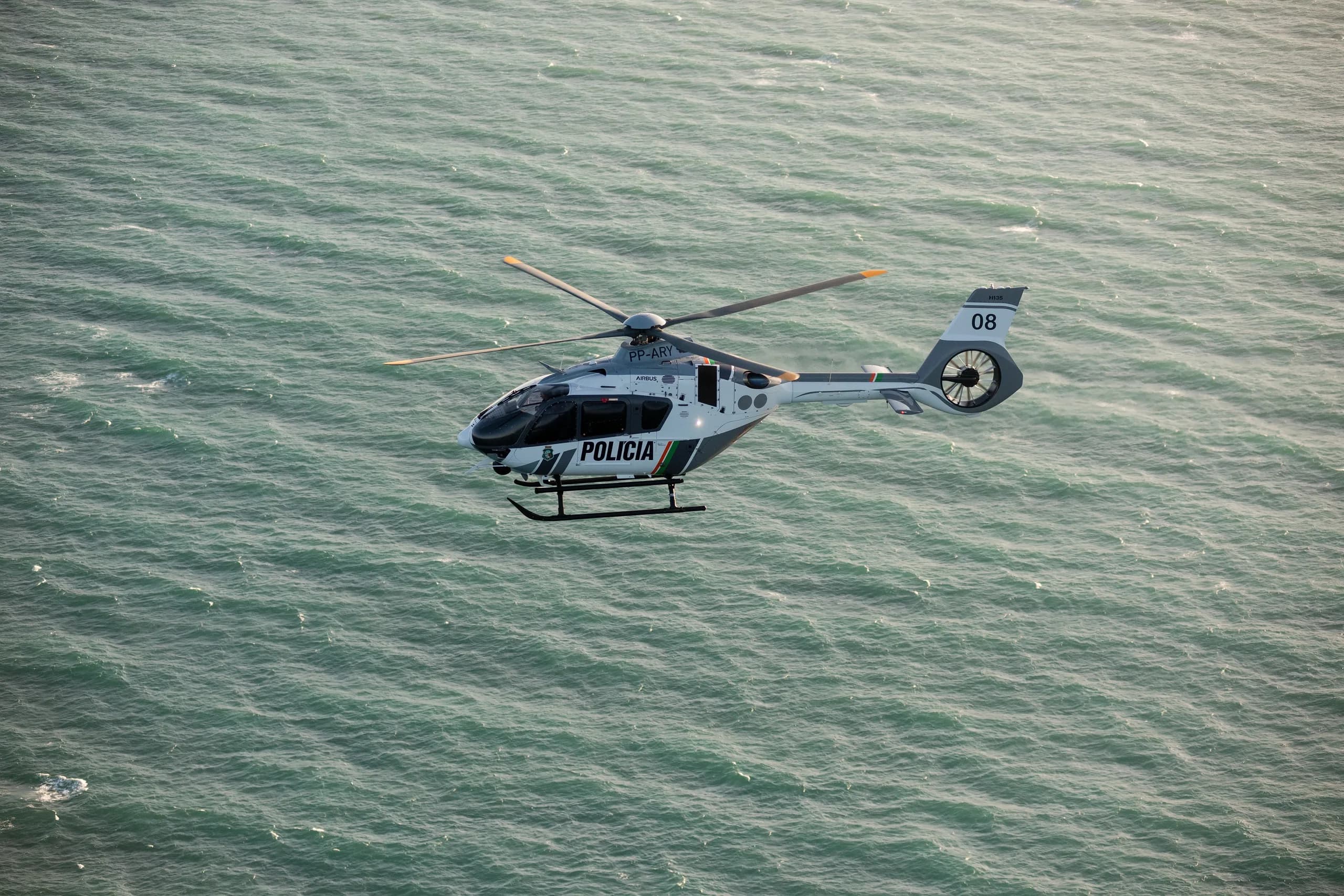 A white and gray Airbus EC135 helicopter with 'POLICIA' markings flying over the ocean.