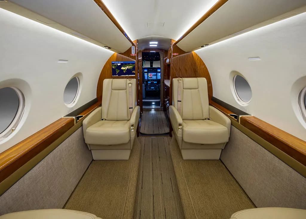 The luxurious interior cabin of the aircraft features beige leather seats and wood paneling, looking towards the cockpit.