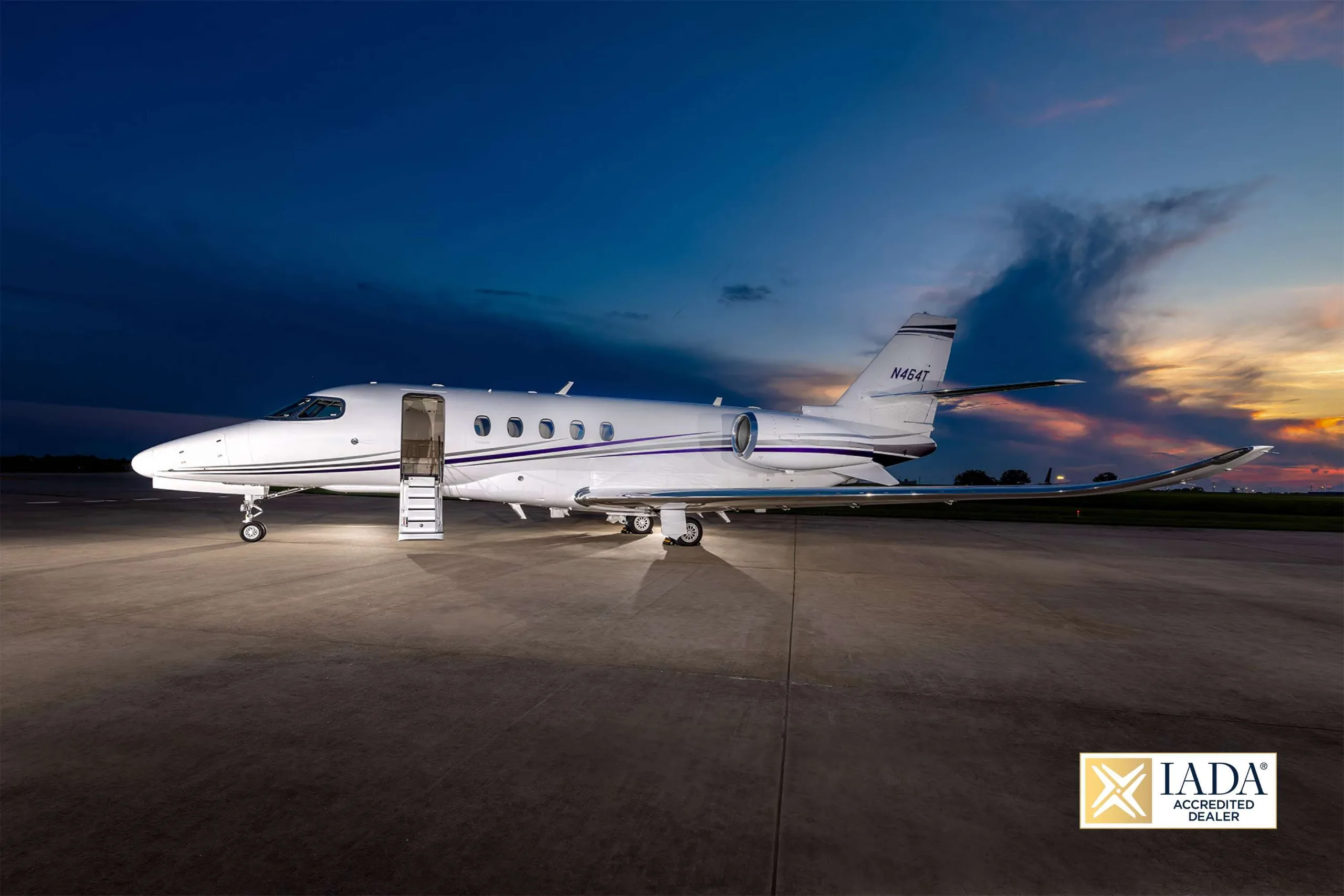 The N464T business jet is parked on an airfield at dusk with its door open, illuminated against a colorful sky.