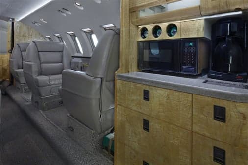 The galley area of a private jet with a microwave, coffee maker, and wooden cabinetry, looking into the cabin.