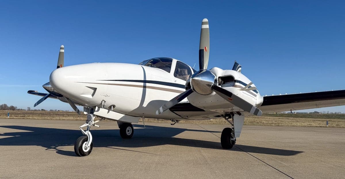 Front-right view of a white twin-engine propeller aircraft with black and gold stripes on an airfield.