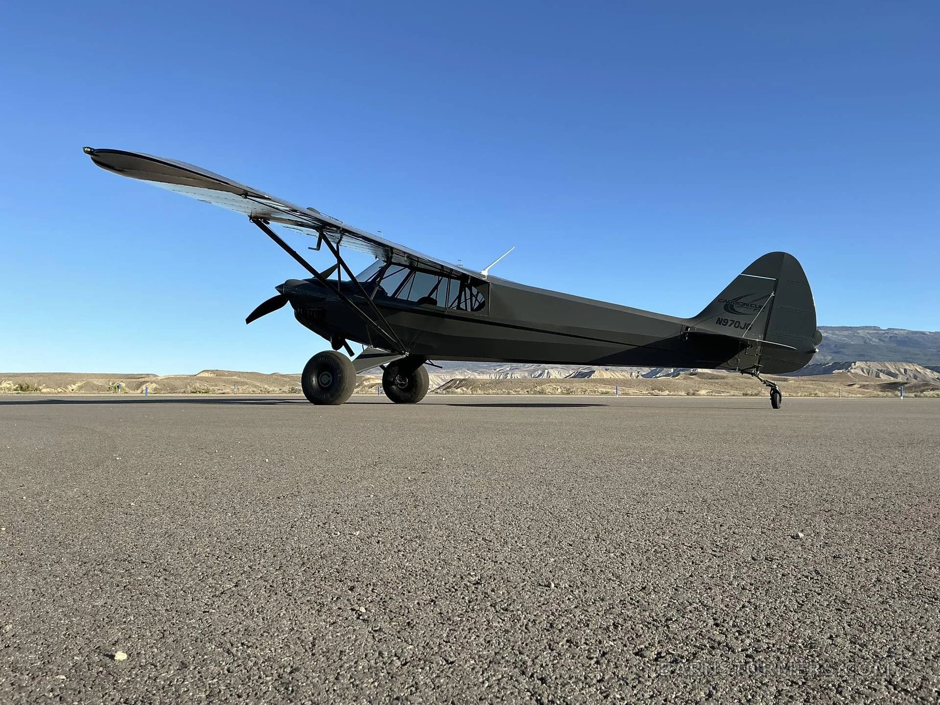 A dark grey Carbon Cub FX3 aircraft with registration N870JH parked on an asphalt runway with mountains in the background.