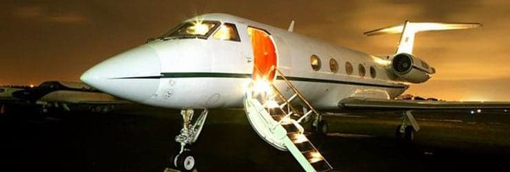 A white business jet with its door open and illuminated stairs extended, parked on a tarmac at night.