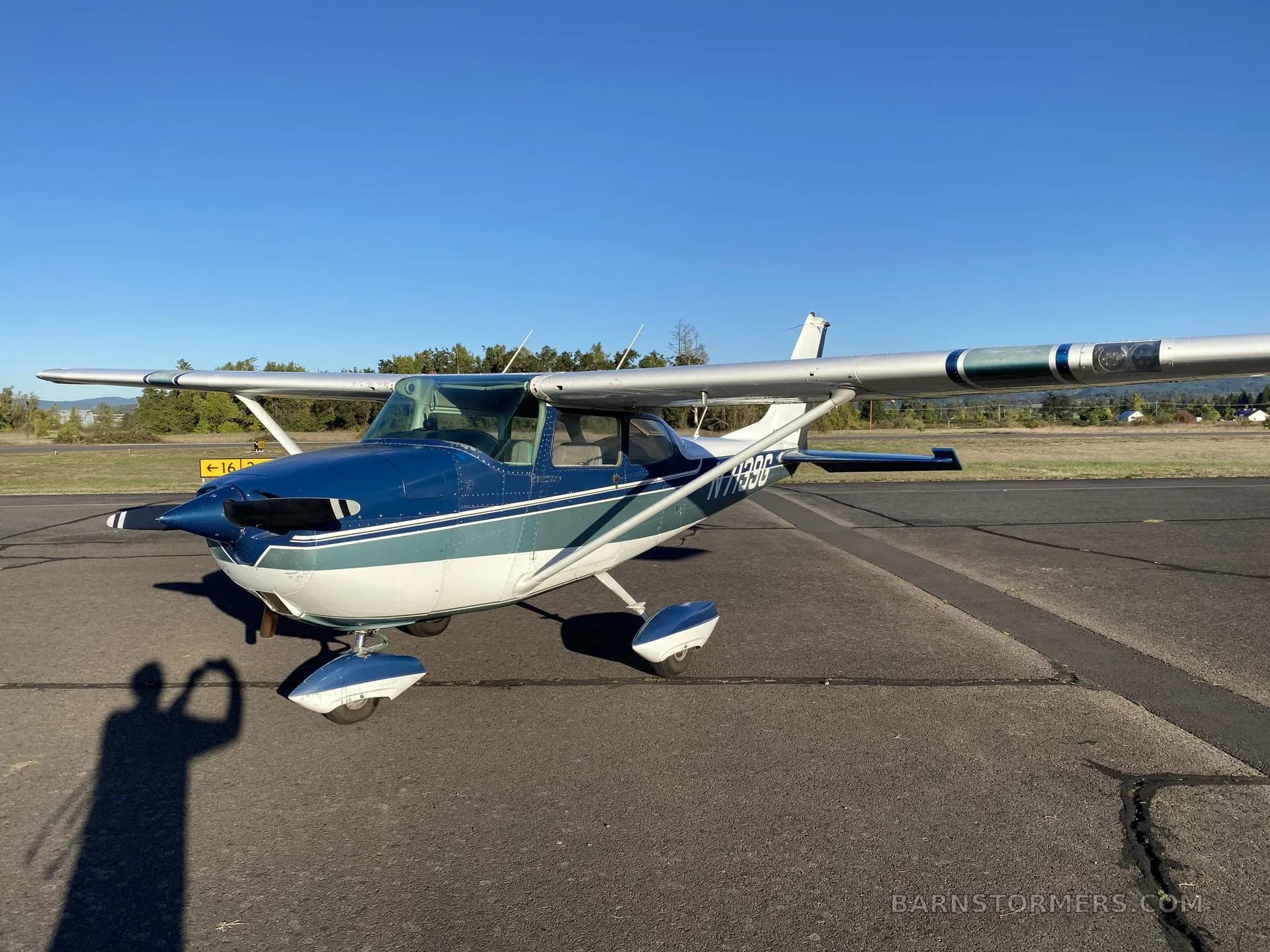 A blue and white Cessna N7199G aircraft is parked on an asphalt ramp, viewed from the front-left, under a clear blue sky.
