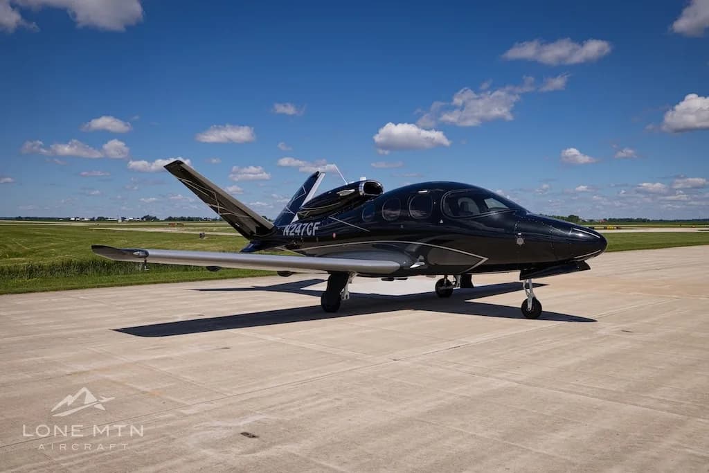 A black Cirrus Vision Jet (N247CF) with silver accents parked on a concrete tarmac under a blue sky with clouds.