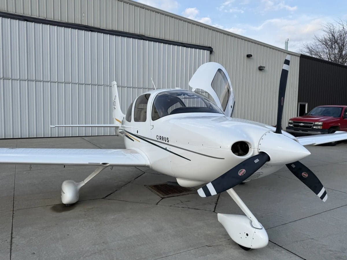 A Cirrus SR20 aircraft is parked outside a hangar with open doors.
