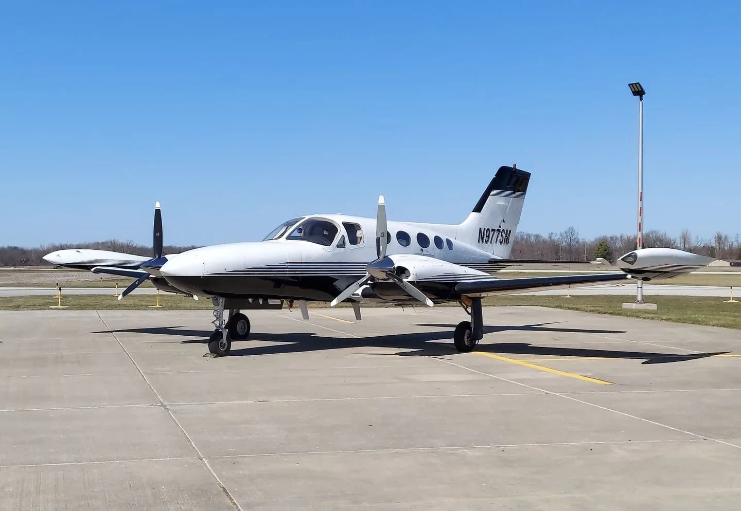 A white and black twin-engine propeller aircraft, N977SM, is parked on a tarmac under a clear blue sky.