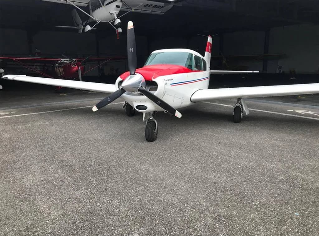 A white single-engine aircraft with a red nose and blue stripes, parked inside a hangar, viewed from the front.