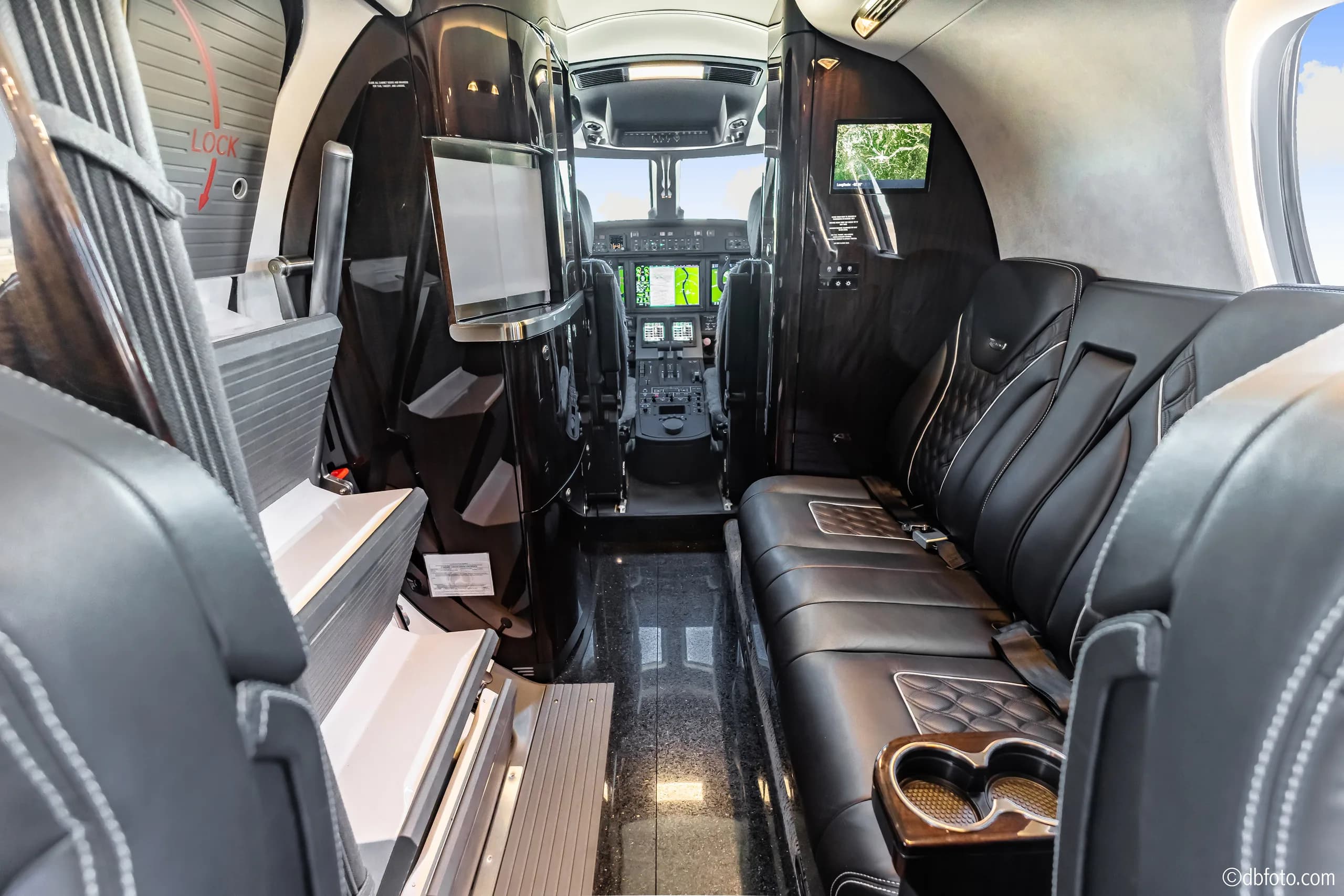 The business jet's cabin looking forward, showing a side couch, entertainment screens, and a glimpse of the cockpit.