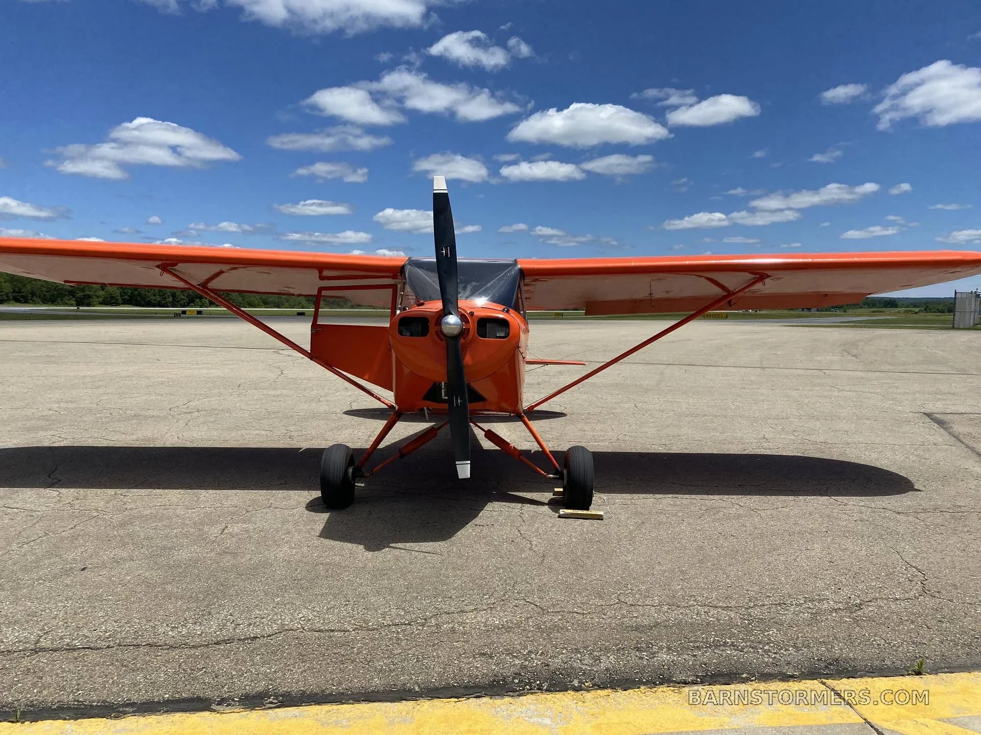 A front view of a bright orange single-engine aircraft with a propeller, parked on an asphalt runway.
