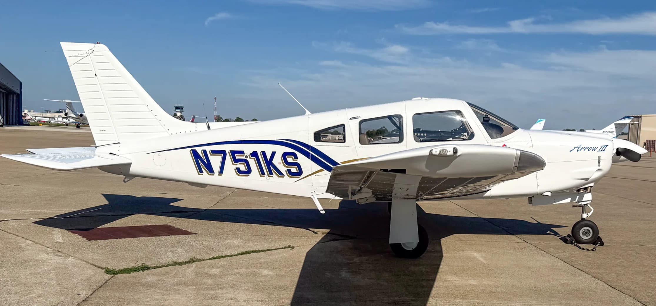 A white Piper Arrow III aircraft with blue and gold stripes, tail number N751KS, seen from the left side on an airport tarmac.