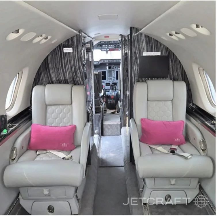The interior of a private jet cabin, looking forward towards the cockpit, with two light grey seats in the foreground.