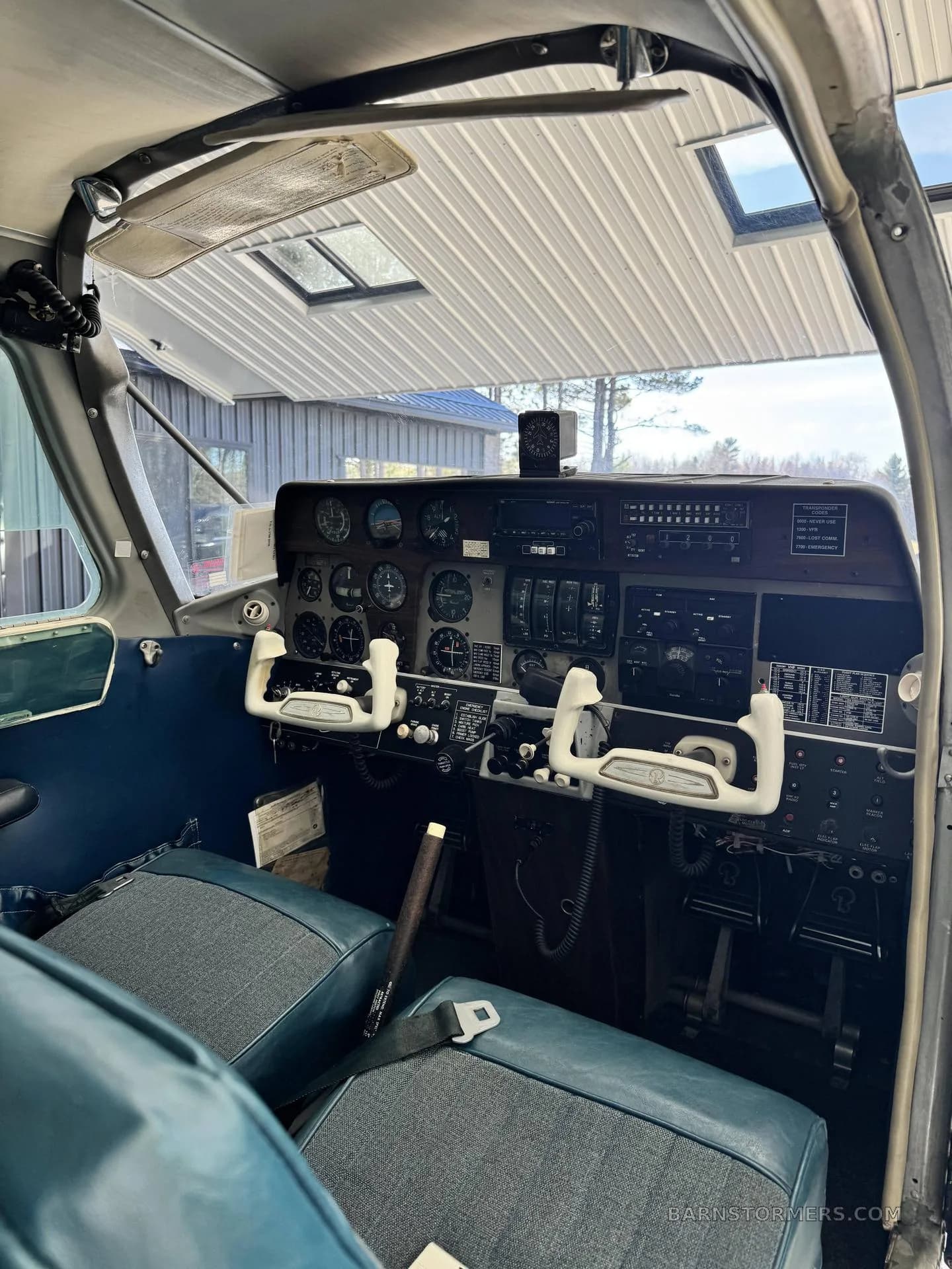The cockpit of a small aircraft, showing the instrument panel with gauges, radios, and two white control yokes.