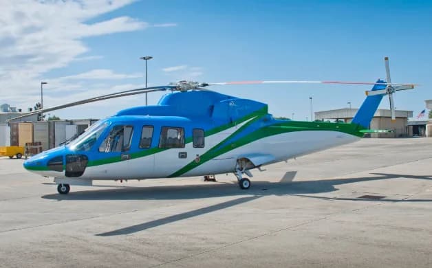 A blue, green, and white helicopter, likely an AgustaWestland AW139, is parked on an airfield tarmac under a partly cloudy sky.