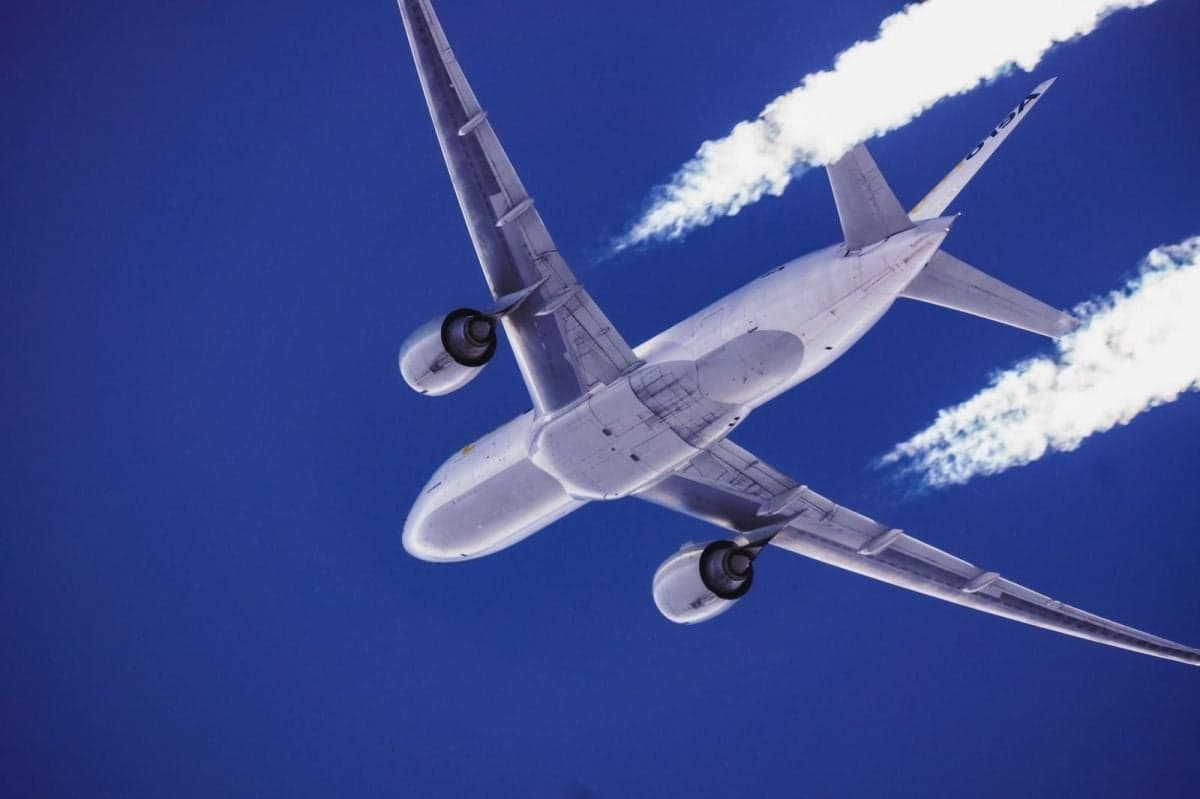 A large passenger jet, possibly a Boeing 777, viewed from below against a deep blue sky, leaving white contrails.
