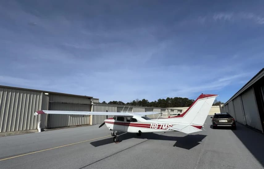 A rear-right view of a white and red Cessna 182 Skylane (N87MS) parked on an airport tarmac with a car in the background.