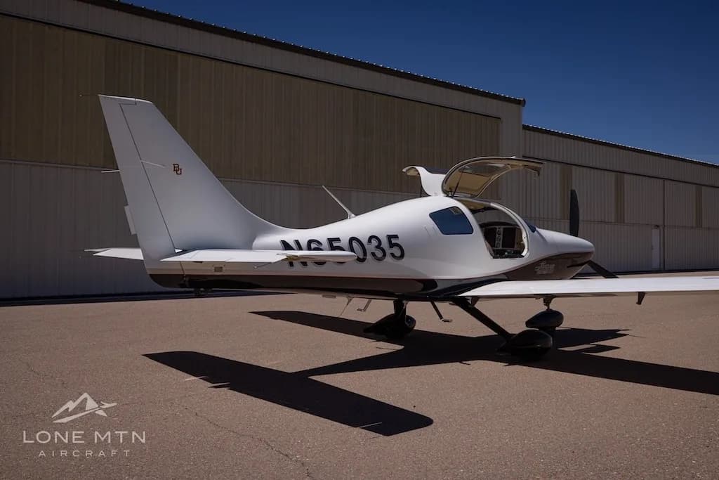 A white and black single-engine aircraft, N65035, with its gull-wing doors open, seen from the rear-right in front of a hangar.
