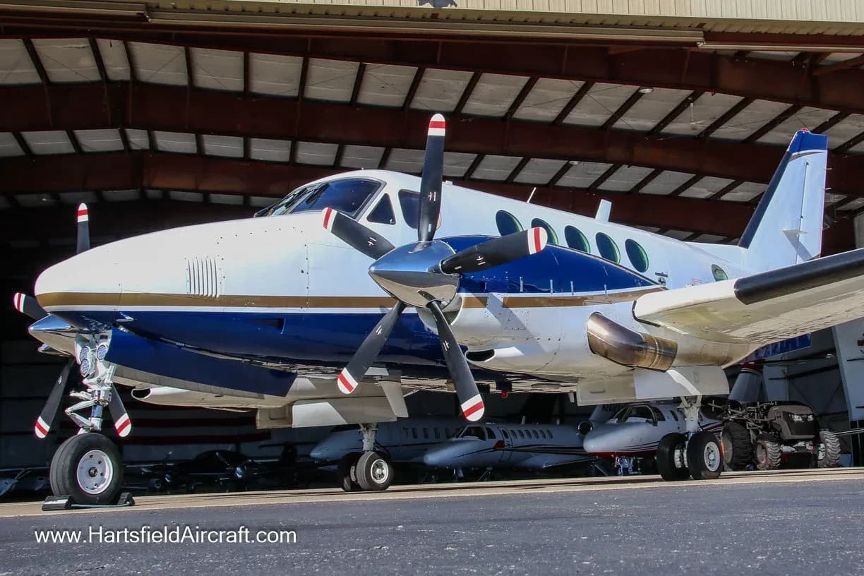 A white and blue twin-turboprop aircraft, likely a King Air, is parked on the tarmac in front of a hangar.